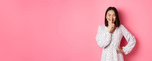 Beautiful Korean woman in trendy dress asking to keep secret, hushing with soft smile and closed eyes, standing over pink background photo