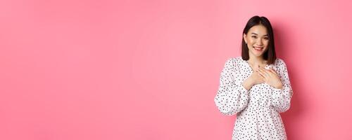 Thankful Korean girl in dress smiling, holding hands on heart and looking grateful at camera, touched with nice gesture, standing over pink background photo