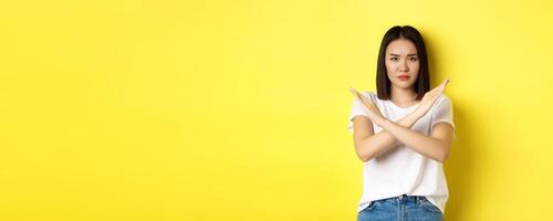 Serious and assertive korean woman showing cross stop gesture, frowning and telling no, prohibit action, disapprove something bad, standing over yellow background photo