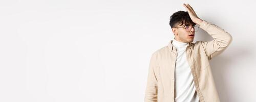 Portrait of annoyed stylish guy facepalm, roll eyes from something stupid, standing on white background tired photo