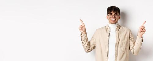 Image of smiling handsome man in glasses pointing fingers sideways, showing advertisements or variants, standing cheerful on white background photo