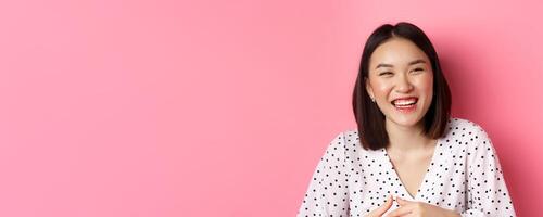 Beauty and lifestyle concept. Close-up of happy asian woman laughing and having fun, standing over pink background photo