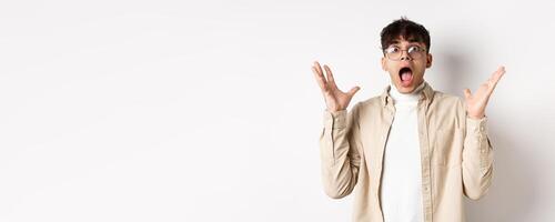 Scared young man in panic, screaming and looking axious, jumping startled and shocked, shaking hands nervously, standing on white background photo