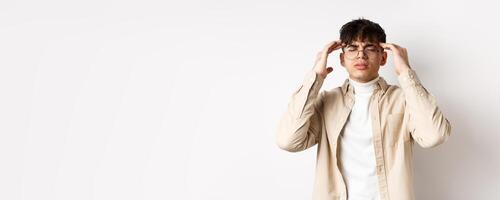 Image of young man in glasses having headache, touching head temples and frowning from painful migraine, feel dizzy, standing on white background photo