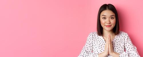 Beauty and lifestyle concept. Close-up of cute asian woman smiling, showing thank you gesture, holding hands in pray, standing over pink background photo