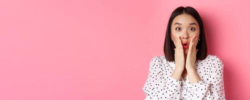 Close-up of surprised and amazed asian woman staring at camera, gasping impressed, holding hands near mouth, standing over pink background photo