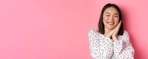 Beauty and lifestyle concept. Close-up of beautiful and dreamy asian woman sleeping on her hands, close eyes and smiling, daydreaming on pink background photo
