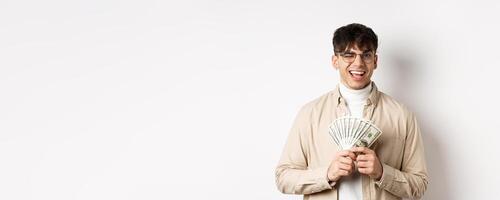 Handsome young man winking and showing dollar bills, holding fan of money banknotes and smiling pleased, making money, standing on white background photo