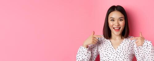Close-up of happy and confident asian woman pointing at herself, smiling self-assured, standing over pink background photo