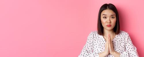Beauty and lifestyle concept. Sad asian woman asking for help, begging with hands in plead gesture, staring at camera, need favour, standing over pink background photo