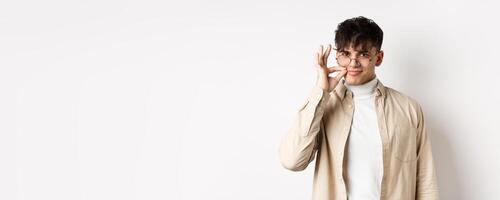 Smiling cunning guy in glasses shut his mouth on zipper, showing seal gesture near lips and smiling devious, keeping a secret, standing on white background photo