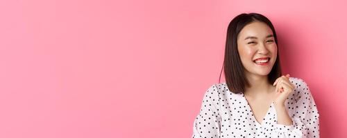Beauty and lifestyle concept. Close-up of happy asian female laughing, looking happy and showing genuine emotions, standing over pink background photo