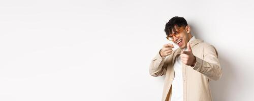 Handsome funny man in glasses smiling, pointing fingers at camera, choosing or inviting you, standing on white background photo