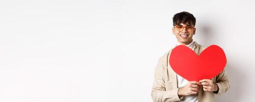 Handsome hipster guy with sunglasses and earring, waiting for true love on Valentines day, holding big red heart and smiling, standing over white background photo