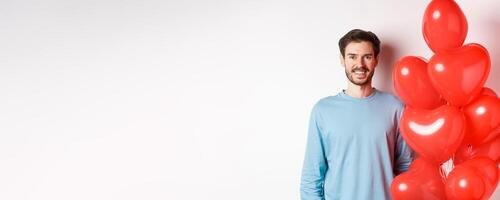 Smiling young man standing with heart balloons and looking happy, celebrating valentines day, bring romantic present to lover, standing over white background photo