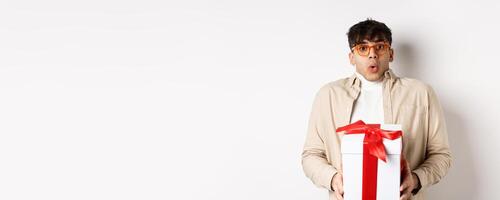 Surprised young man say wow and holding gift box, receive present on holiday, standing amused on white background photo
