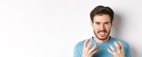 Close up of angry young man with beard, shaking hands mad, squeeze teeth and frowning furious, standing outraged over white background photo