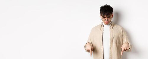 Confused frowning guy in glasses looking, pointing fingers down at something strange, standing puzzled on white background photo