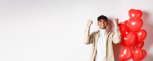 Valentines day. Satisfied young man saying yes, triumphing and celebrating on lovers date, making fist pump and smiling pleased, standing near heart balloons on white background photo