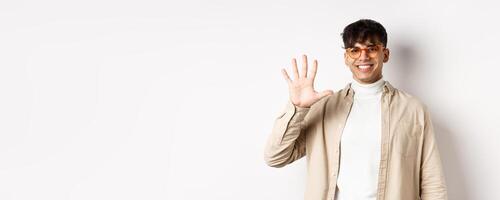 Modern guy in glasses and stylish outfit, showing five fingers number and smiling, making order, standing on white background photo