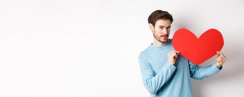 Valentines day. Handsome and romantic man holding big red valentine heart cutout, looking seductive at camera, making love confession, white background photo