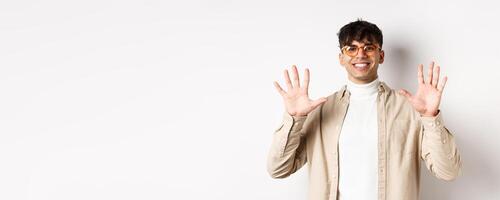 Modern guy in glasses and stylish outfit, showing ten fingers number and smiling, standing on white background photo