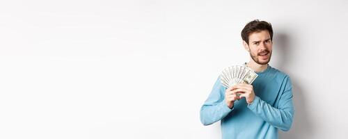 Handsome young man showing money and looking pleased. Guy dancing with dollars, earn income, standing over white background photo