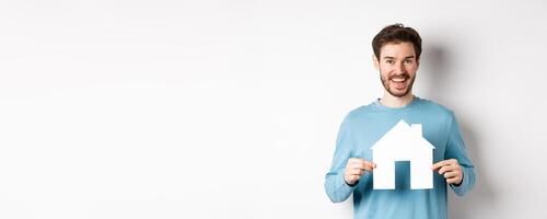 Real estate and insurance concept. Handsome modern man buying property, smiling and showing paper house cutout, standing over white background photo