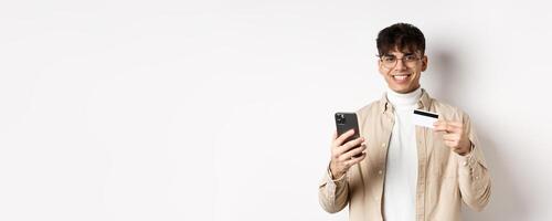 Portrait of natural young man in glasses paying in internet, showing smartphone and plastic credit card, standing on white background photo