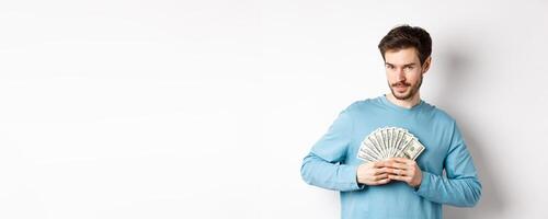 Confident bearded man smiling, showing dollars and looking at camera. Handsome guy holding money on white background photo