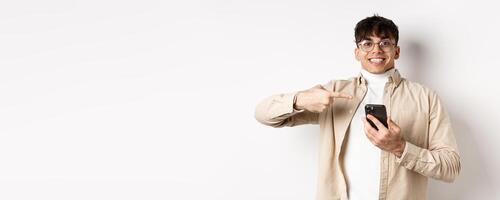 Technology and online shopping concept. Cheerful young man in glasses showing on mobile phone, pointing at smartphone screen and smiling excited, white background photo