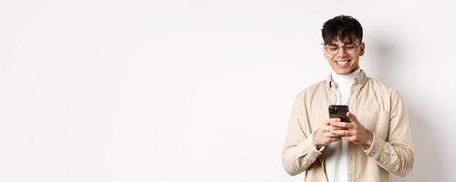 Real people. Natural young man reading message on mobile phone, smiling and looking at smartphone screen, standing on white background photo