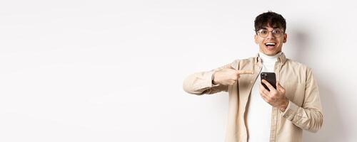 Technology and online shopping concept. Happy hipster guy pointing finger at smartphone screen and smiling, checking out app promo, standing on white background photo