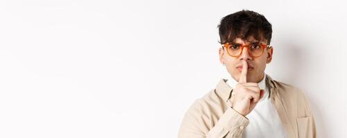 Close-up of grumpy young guy in glasses frowning and shushing, make taboo gesture with finger shuting lips, tell to be quiet, standing angry on white background photo