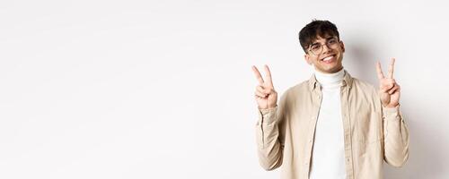 Handsome hipster guy in glasses, smiling with white teeth, showing peace or victory gesture, standing upbeat on white background photo