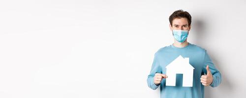 Covid and real estate concept. Smiling agency client showing thumb up and paper house cutout, wearing medical mask, approve good deal photo