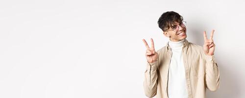 Real people. Handsome hipster guy in glasses showing peace gestures and smiling cute, standing on white background photo