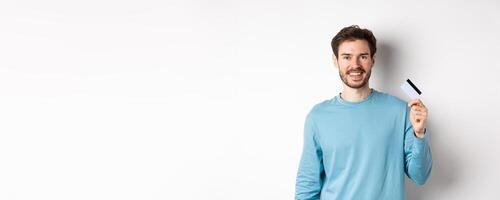 Smiling male bank client showing plastic credit card with happy face, standing over white background photo