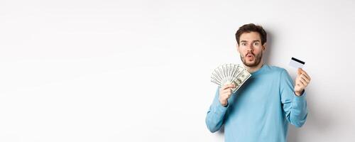 Surprised young man showing money in dollars and plastic credit card, gasping and saying wow with amazed face, standing on white background photo