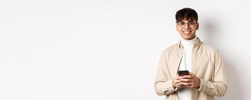Real people. Handsome guy in glasses using mobile phone and smiling happy at camera, standing on white background photo