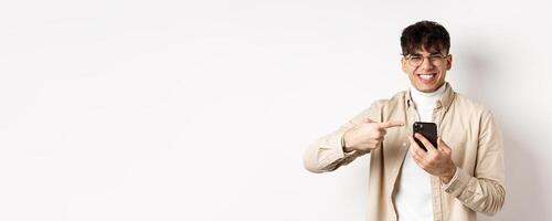 Technology and online shopping concept. Portrait of natural man in glasses laughing and pointing finger at smartphone, showing promo offer on screen, standing white background photo