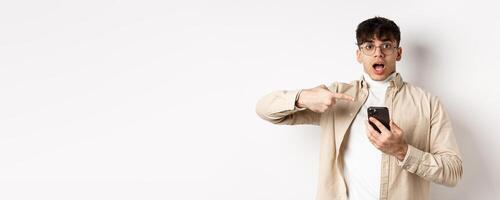 Real people. Amazed young guy checking out online offer on mobile phone, pointing at smartphone screen with excited face, standing on white background photo