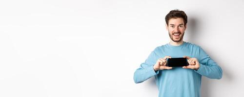 E-commerce and shopping concept. Smiling cheerful man showing empty smartphone screen in horizontal position, looking happy at camera, white background photo