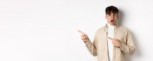 Amazed young man in glasses drop jaw, saying wow, pointing and looking left at awesome promo offer, stare with disbelief and impressed face, white background photo