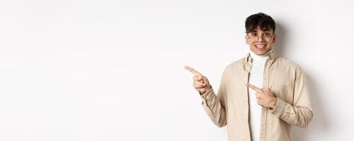 Cheerful handsome man in glasses smiling, pointing fingers left at logo, showing advertisement with excited face, standing on white background photo