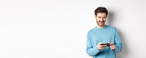 Image of handsome young man playing mobile game, holding smartphone horizontally and smiling at camera pleased, standing over white background photo