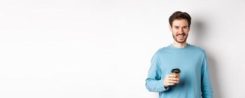 Cheerful man drinking coffee from cafe take-away and smiling, standing with paper cup in white background photo