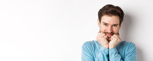 Cute caucasian man looking at something with admiration and desire, touching face with silly smile, standing over white background photo