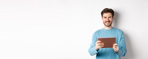 Handsome young man holding digital tablet and smiling, networking with device, standing over white background photo