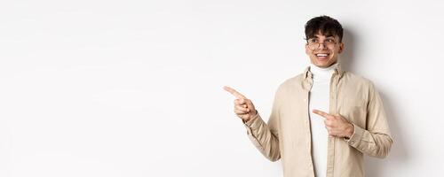 Excited and enthusiastic young man in glasses, pointing fingers and looking left with happy smile, standing in awe on white background photo